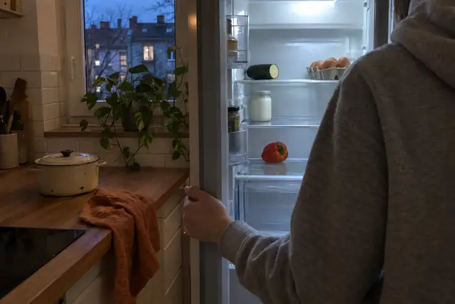 Eine Person im grauen Hoodie steht am Abend vor dem offenen, halb leeren Kühlschrank.