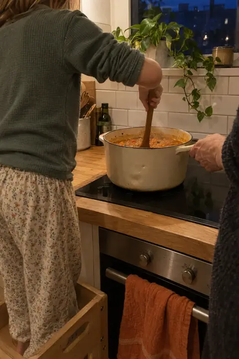 Ein Kind steht auf einem Tritthocker am Herd und rührt die Linsensuppe, eine erwachsene Hand hält den Topf.