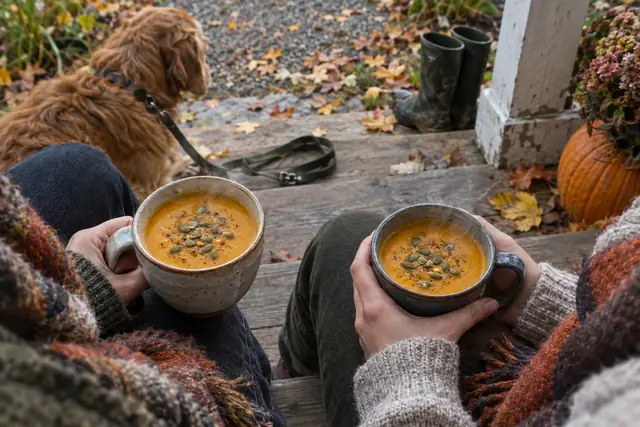 Zwei Personen sitzen auf einer Holzstufe vor dem Haus und halten Becher mit Kürbissuppe in den Händen.